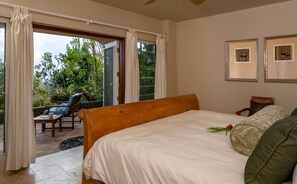 A bedroom with a bed, a chair, a table, and a view of greenery outside. - Mango Bay: Amazing North Shore Views! (St. John)