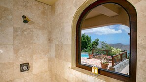 Une salle de bain avec douche, une fenêtre donnant sur une piscine et un espace extérieur, ainsi qu’une petite étagère avec des bouteilles.