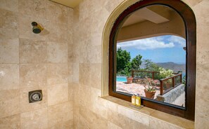 A bathroom with a shower, a window with a view of a pool and outdoor area, and a small shelf with bottles. - Mango Bay: Amazing North Shore Views! (St. John)