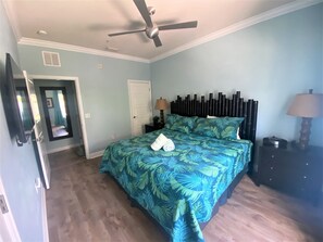 A bedroom with a bed, a ceiling fan, a nightstand with a lamp, and a door leading to another room.