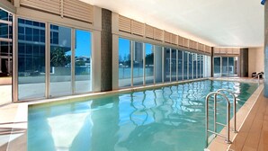 A large indoor swimming pool with clear glass walls, a tiled floor, and a metal ladder.