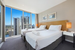 Ein Hotelzimmer mit zwei Betten, einem Nachttisch mit Lampe und großen Fenstern mit Blick auf die Stadt.