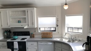 A compact kitchen with white cabinets, a stove, a dishwasher, and a sink. There are two windows with blinds, a ceiling light, and a wooden rack on the countertop. - The Moon House. Come mingle with the stars. (Hotchkiss)