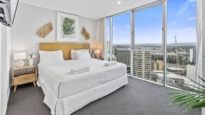 Habitación de hotel con una cama grande, una mesita de noche, una lámpara y vistas de la ciudad a través de ventanales de gran altura.