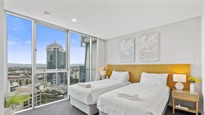 Habitación de hotel con dos camas, un ventanal grande con vista a la ciudad, cabecera de madera y mesita de noche con lámpara.