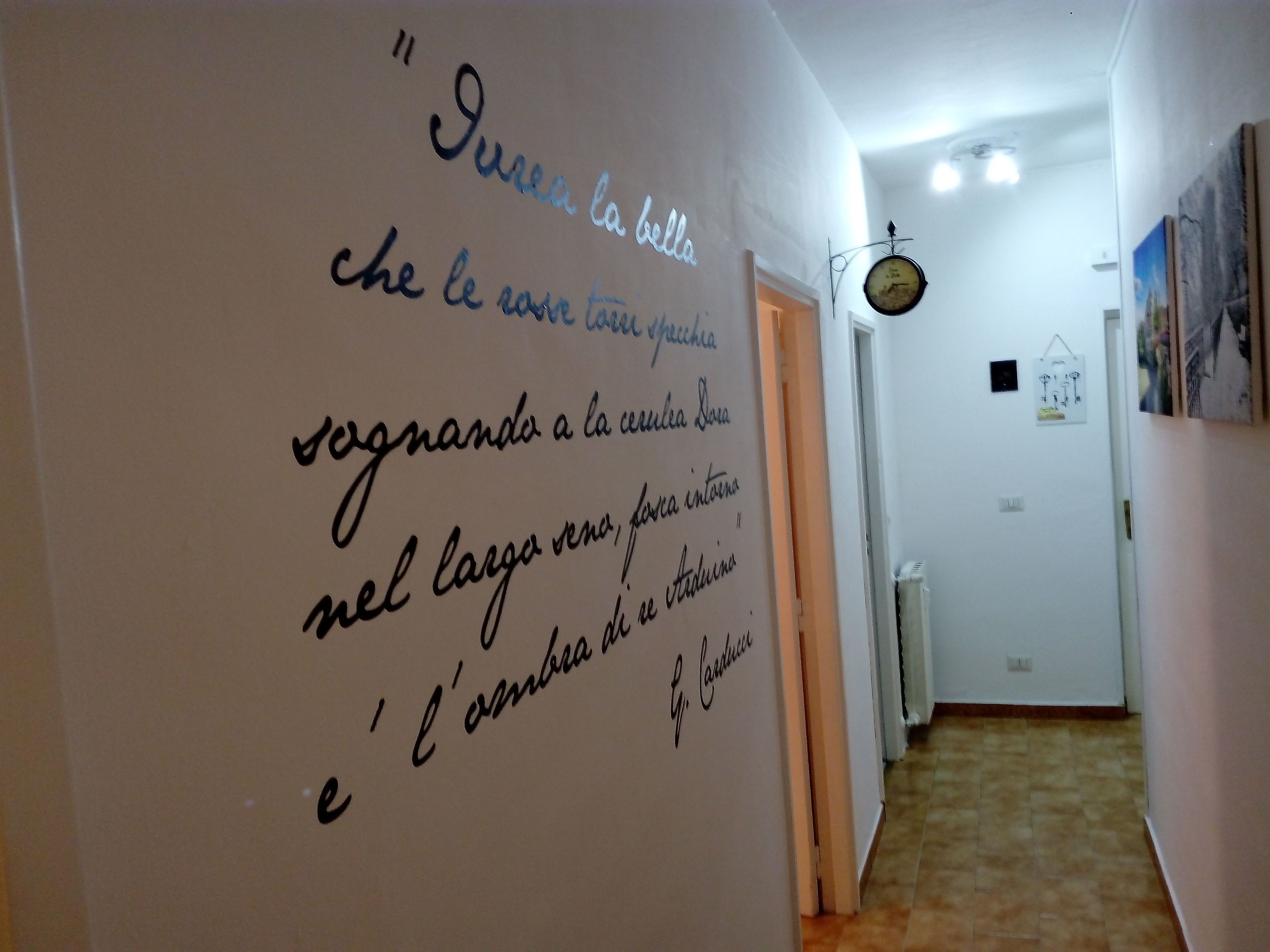 Un corridoio con una parete bianca su cui è scritta a mano dell'iscrizione in una lingua straniera. Si vede una porta alla fine del corridoio e un orologio appeso al muro.