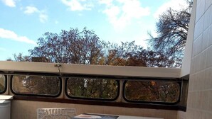 Un petit balcon avec deux chaises blanches et une table, un sol carrelé et une vue sur les arbres et le ciel, visible à travers une fenêtre.