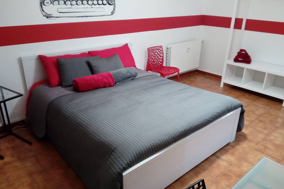 A neatly made bed with grey and red bedding, a red chair, and a white shelf unit in a room with a red and white striped wall.
