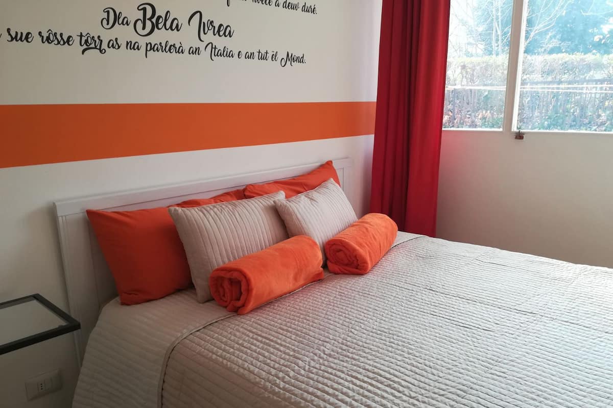 A neatly made bed with orange and white bedding, a red curtain, and a wall with a partial view of a sign in a foreign language.