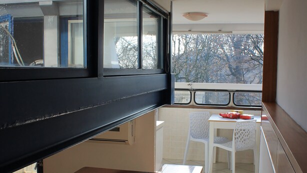 Un balcon étroit avec une table et des chaises, une fenêtre donnant sur un immeuble et un sol carrelé.