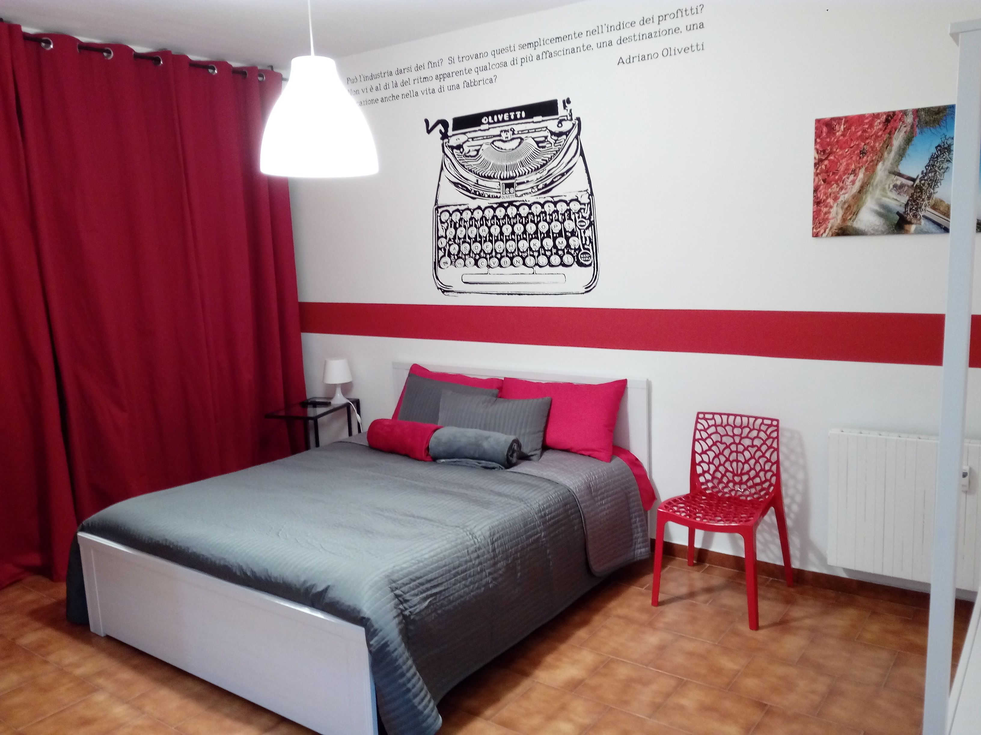 A bedroom with a bed, red curtains, a red chair, a wall with a typewriter illustration, and a radiator.