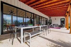 Un'area pranzo all'aperto con un tavolo bianco e sedie in metallo, un soffitto in legno e ampie finestre.