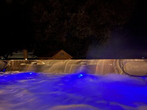 A hot tub with water jets and blue lighting, surrounded by trees and a building in the background.