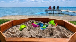 A wooden sandbox with various beach toys, a wooden deck with chairs, and a view of the sea. - Lake Simcoe Waterfront Retreat | Private Dock, Sandy Beach & Hot Tub (Georgina)