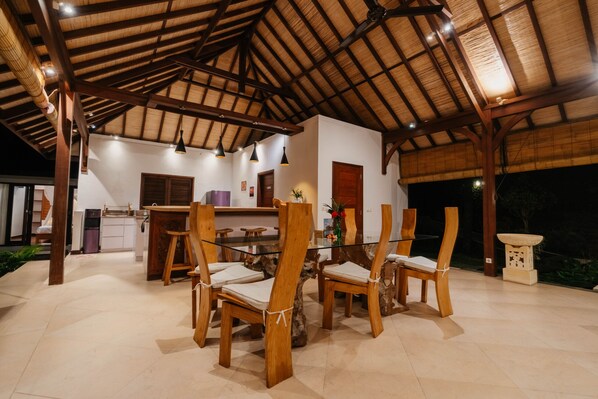 An outdoor dining area with wooden chairs and a glass table, under a wooden roof structure.