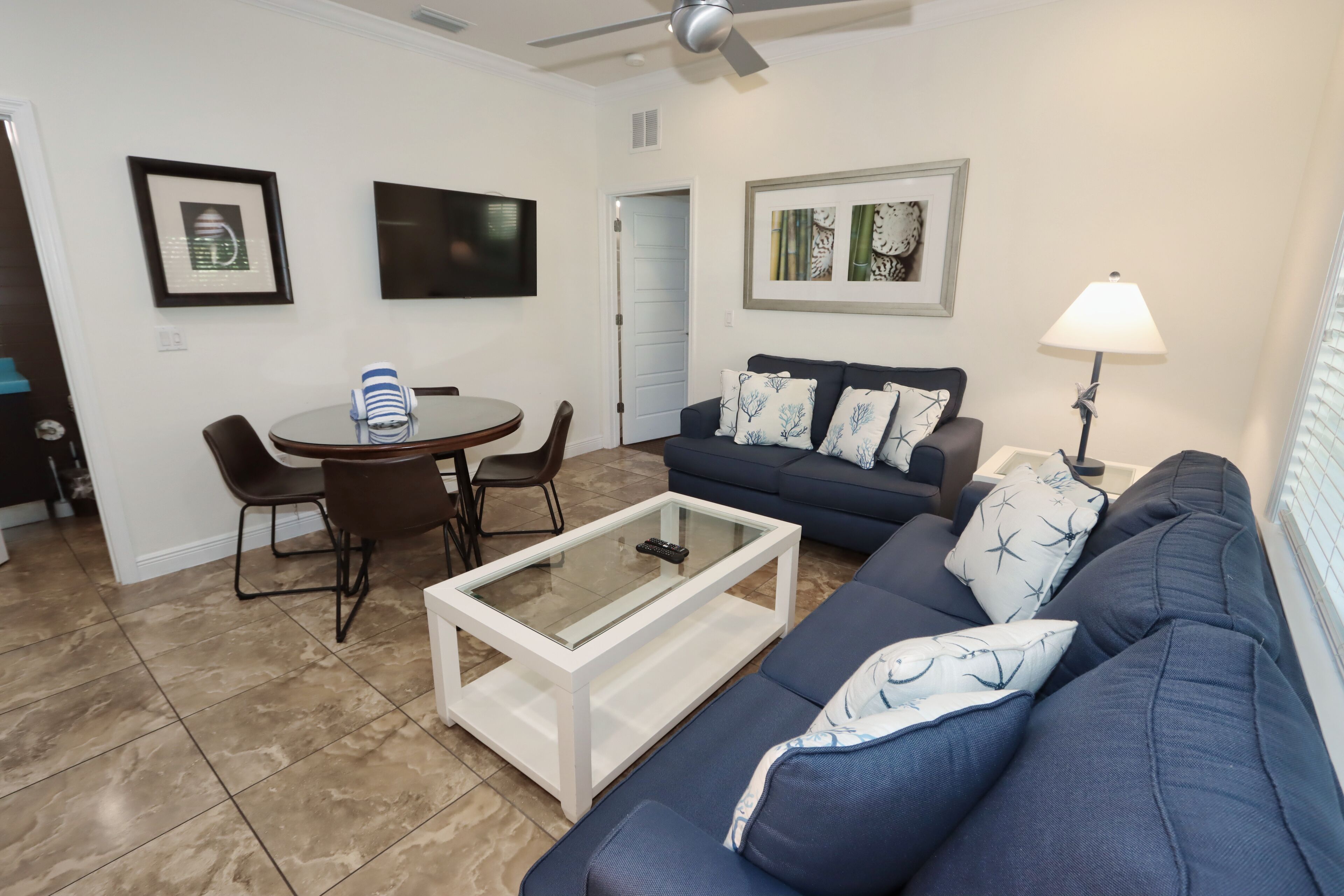 Una sala de estar con un sofá azul oscuro, una mesa de centro de vidrio, una mesa de comedor con sillas y un televisor montado en la pared.