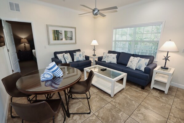A living room with a dark blue sofa, two side tables with lamps, a glass coffee table, and a dining table with chairs. - Tropical Waters & Sunsets–Beach Club Siesta 106B RVA (Sarasota)