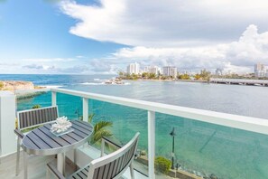 Balcón con mesa y sillas con vista a un mar azul cristalino y un paisaje urbano.
