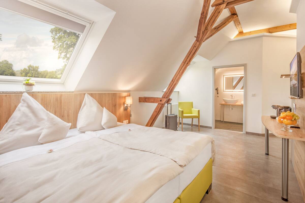 A bedroom with a sloped ceiling, a bed, a bedside table with fruit, a chair, and a bathroom visible through an open door.