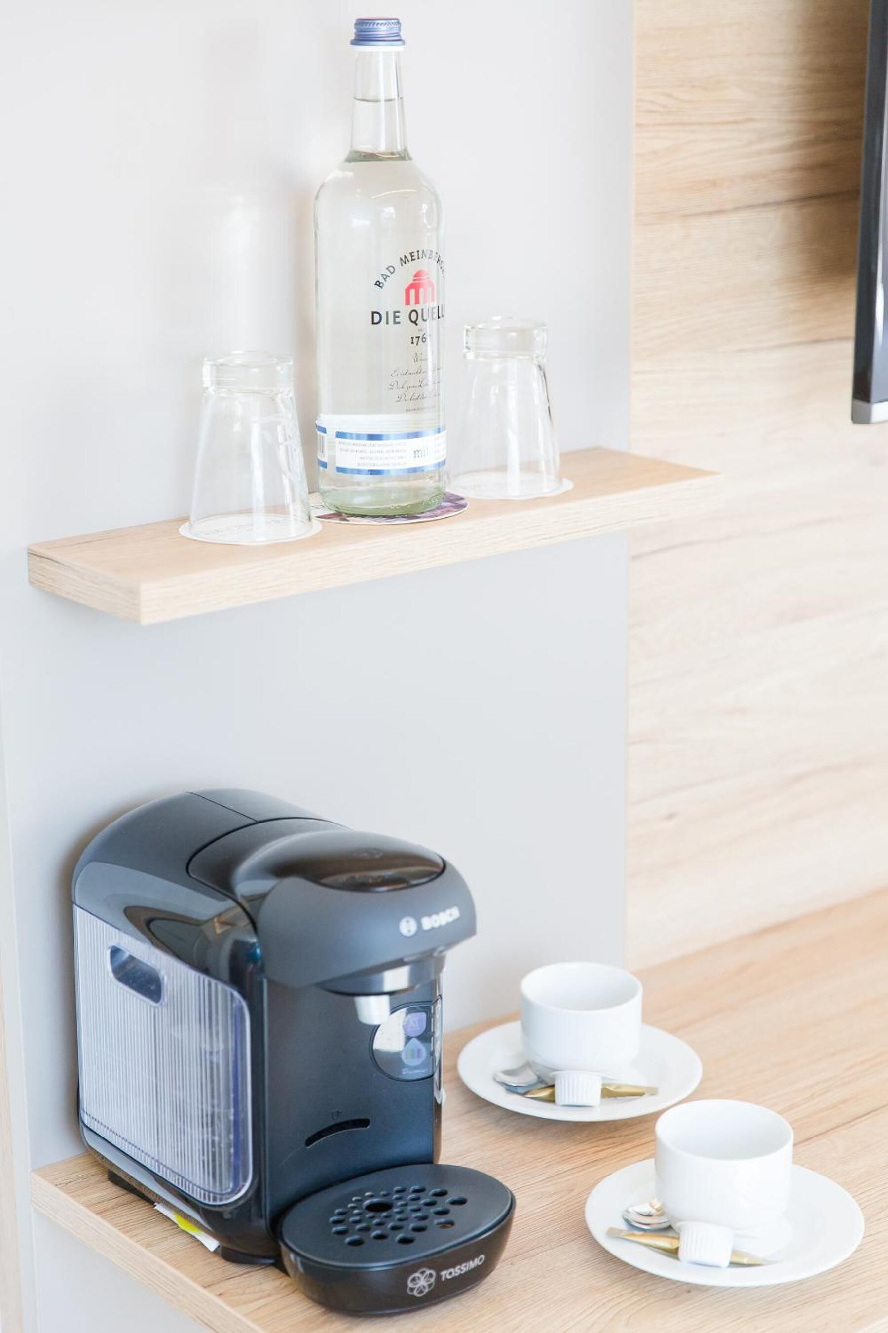Una cafetera sobre un estante de madera con dos tazas blancas y una botella de licor en un estante más pequeño arriba.