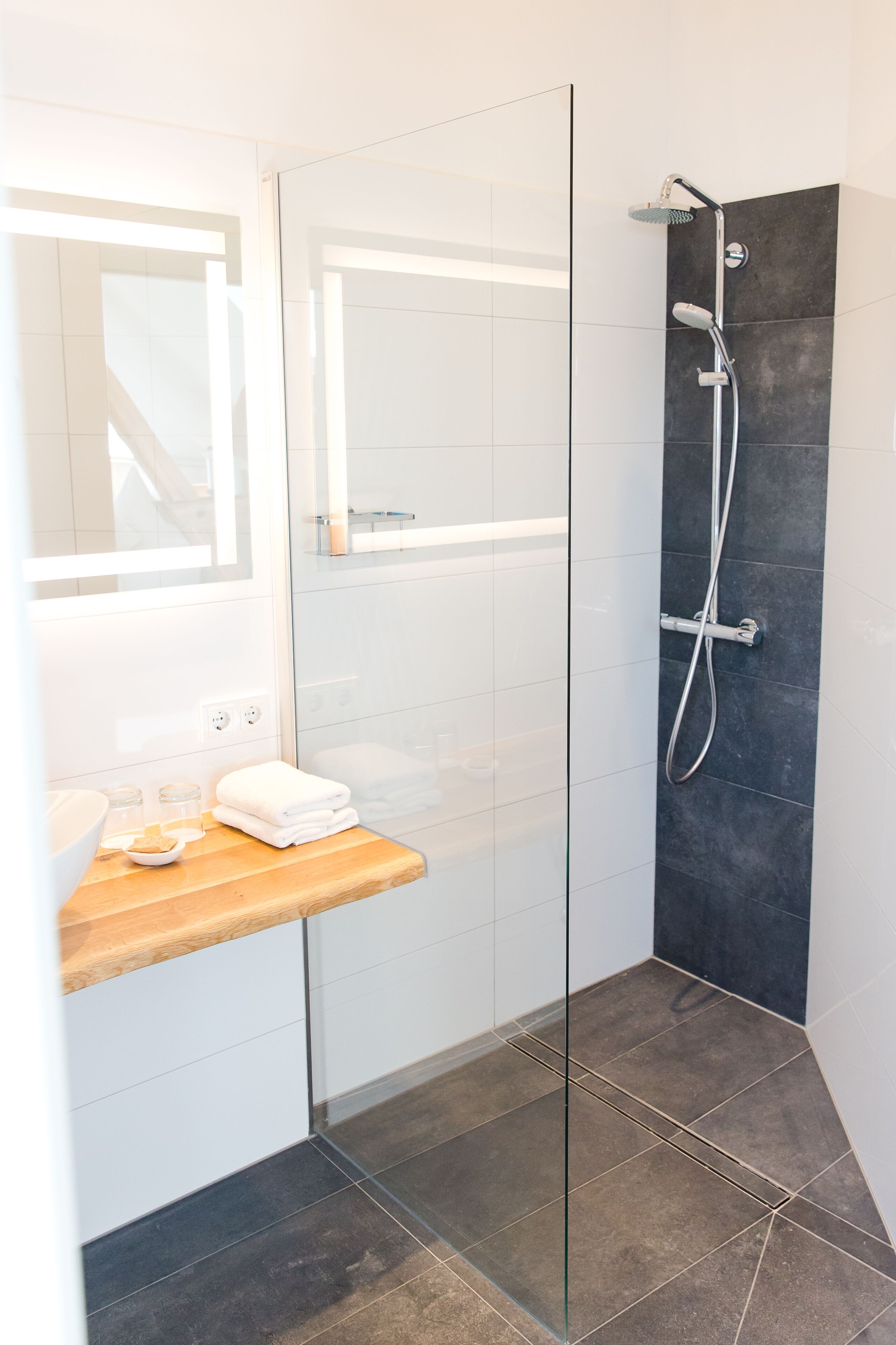 A modern bathroom with a glass shower enclosure, a wooden vanity, and a dark tiled shower wall.