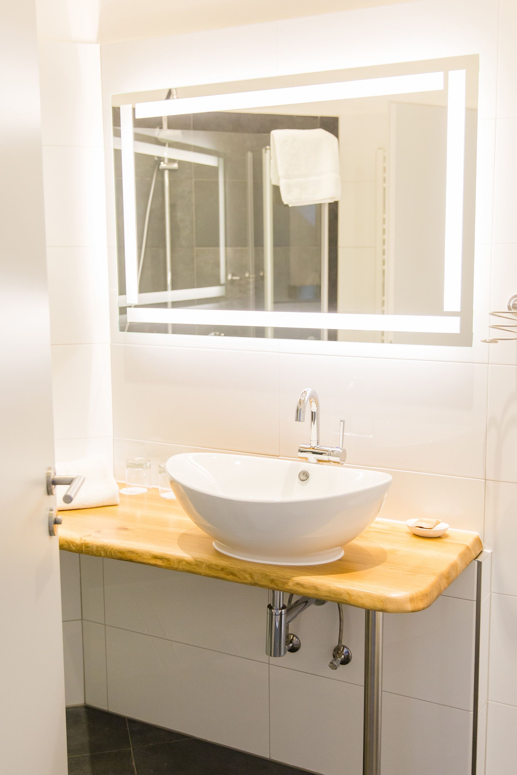 Ein Badezimmer mit einem weißen Waschbecken, einem Spiegel darüber und einer durch die Glastür einsehbarer Dusche.