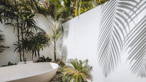 Un baño moderno con una bañera blanca grande, rodeado de exuberante follaje tropical y una pared blanca.