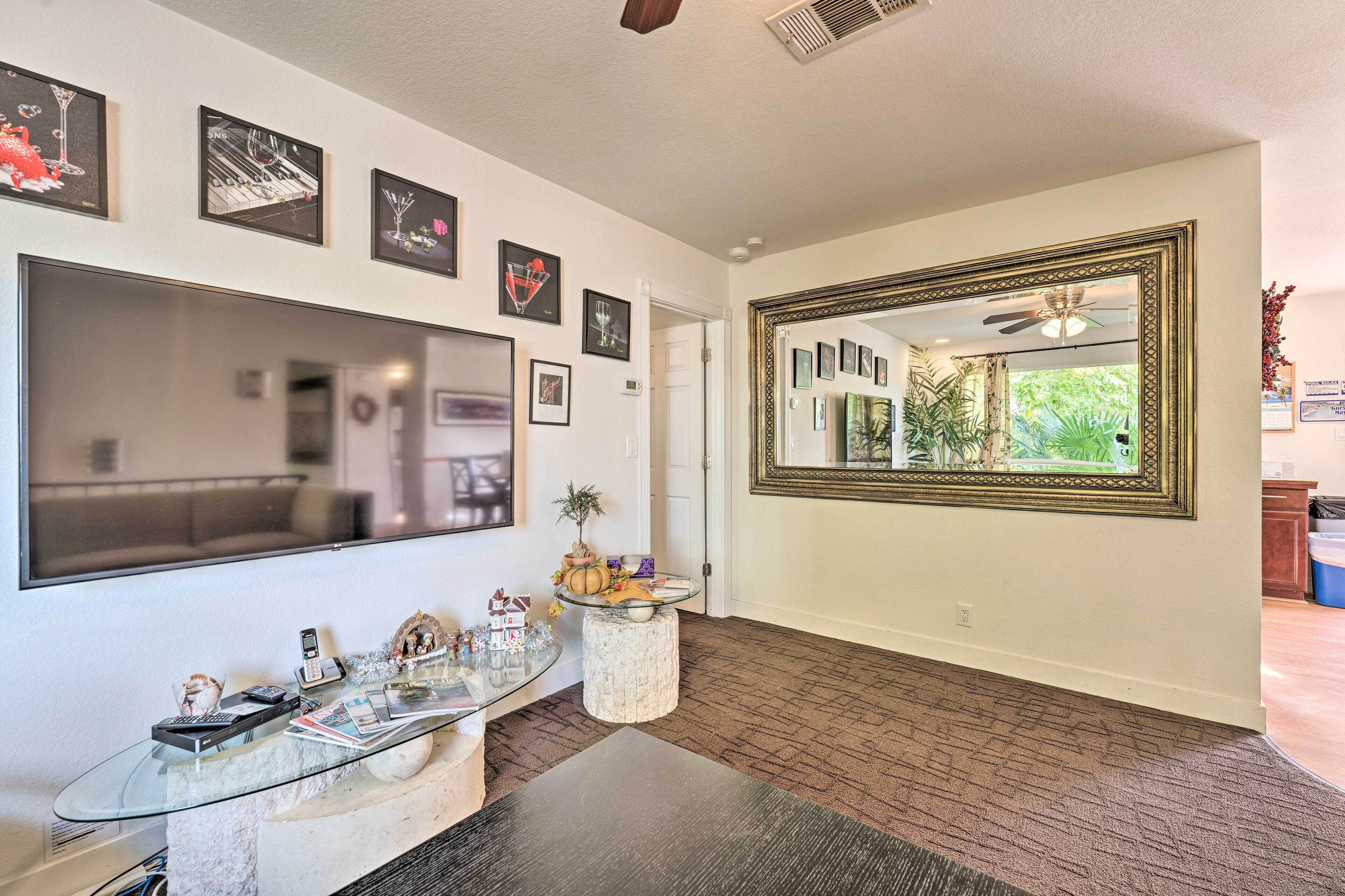 A modern living room with a large flat-screen TV, a glass coffee table, and a decorative mirror.