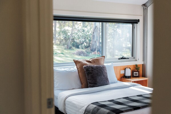 Un dormitorio con un ventanal grande, una cama con ropa de cama blanca, una mesita de noche con una lámpara y vistas a árboles desde el exterior.