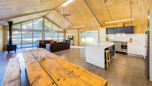 A spacious living area with a wooden ceiling, large windows, a central wooden table, and a modern kitchen with white cabinets and stainless steel appliances. - Nine Steps - private estate on 29 acres with stunning views of Mount Buffalo (Porepunkah)