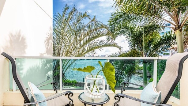 Un balcón con dos sillas, una mesita pequeña, una cesta decorativa blanca y vistas al mar y a las palmeras.