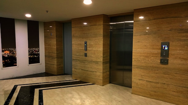 Un ascensor moderno con pantallas digitales, suelo de mármol con líneas negras y vistas a la ciudad a través de un ventanal.