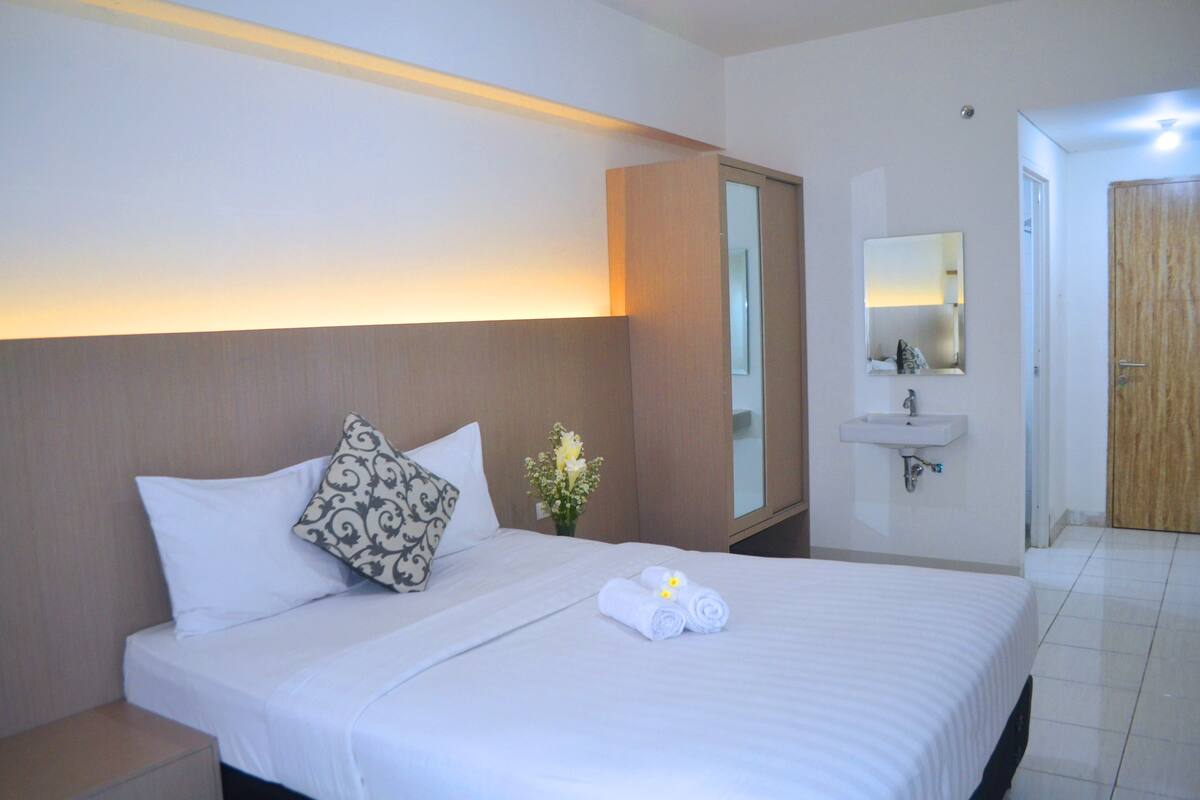 A hotel room with a bed, a nightstand, a mirror, a sink, and a door.