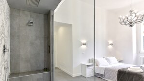 A modern bedroom with a glass shower enclosure, a bed with white linens, and a chandelier.