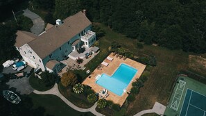 An aerial view of a large house with a swimming pool, a tennis court, and a well-maintained lawn. - Luxurious 7BR Estate Includes - Tennis Crt, Pool, Hot Tub, Home Theater, & Gym! (West Barnstable)