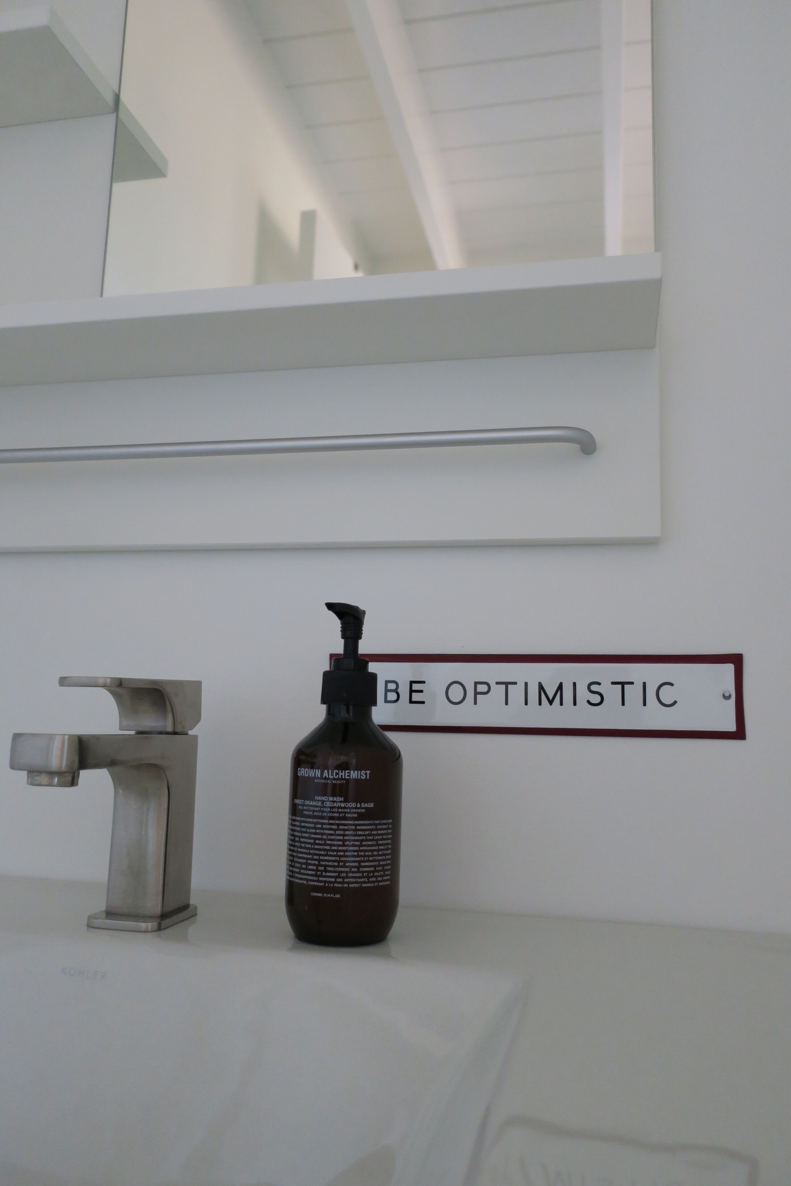 Une salle de bain équipée d’une barre d’appui, d’un robinet et d’un flacon de savon de la marque Crown Alchemist. Une pancarte au-dessus indique : « SOYEZ OPTIMISTE ».