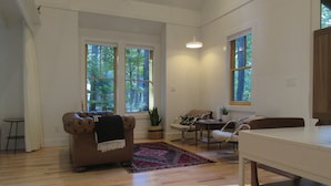 Amplia sala de estar con pisos de madera, un sofá de cuero marrón, una pequeña mesa redonda y dos sillas. Amplias ventanas ofrecen luz natural y vistas a árboles.