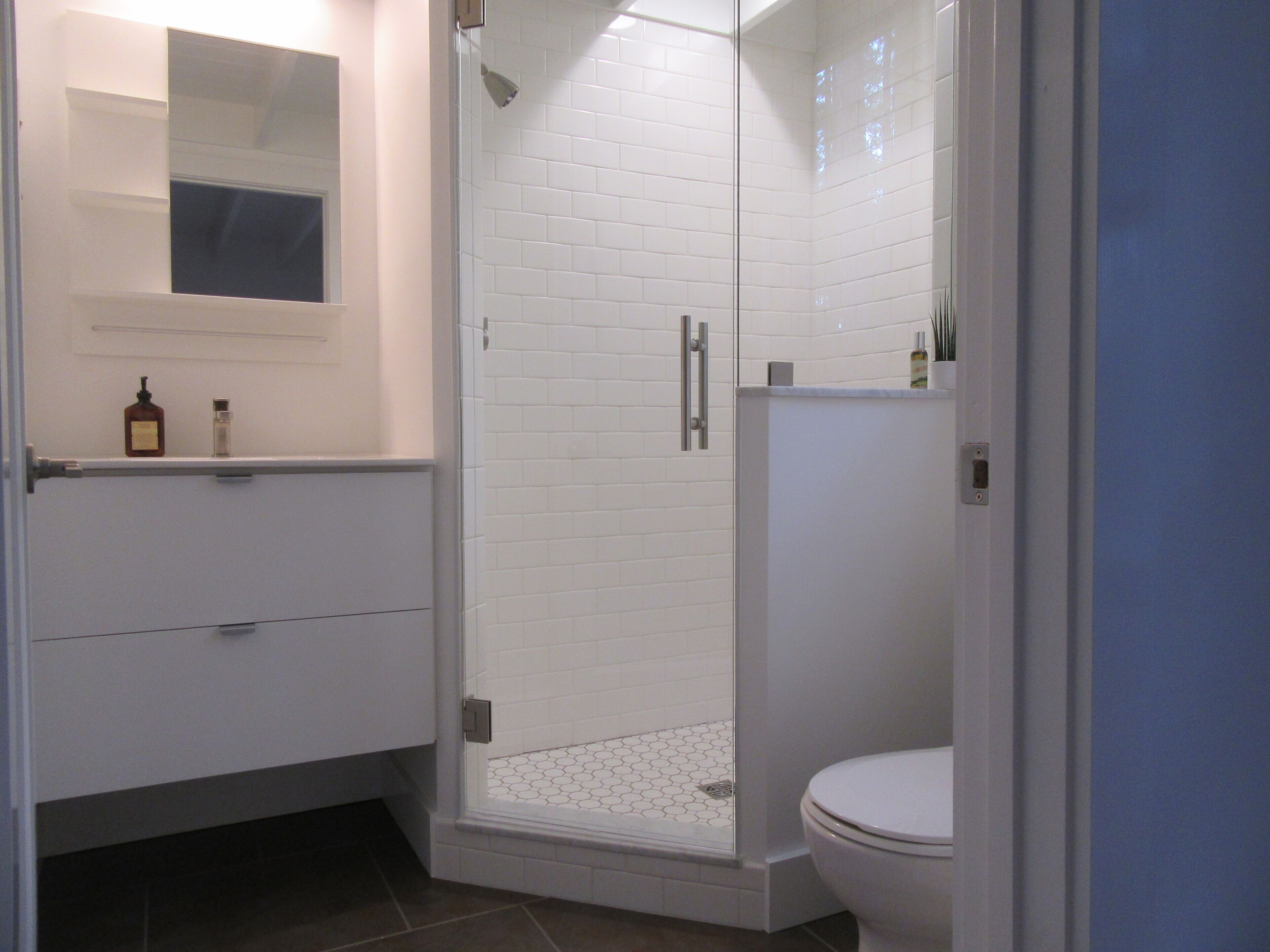 Un baño moderno con ducha de vidrio, azulejos blancos, inodoro y espejo sobre el lavamanos.