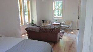 Una habitación amplia con una cama grande, un sofá marrón, dos sillones, una mesita y un ventanal que proporciona luz natural.