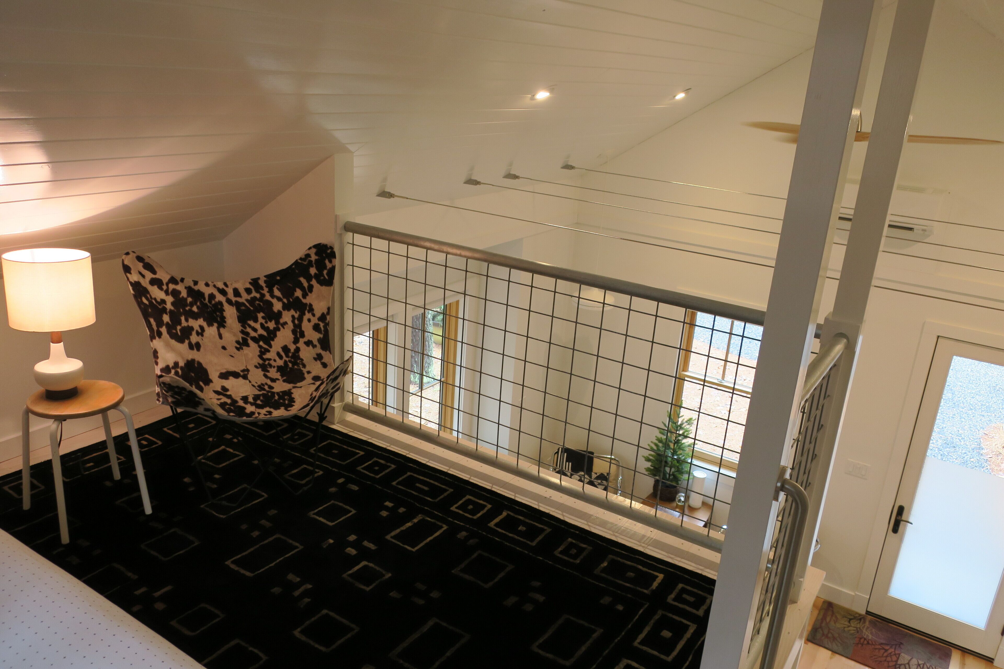 Une pièce avec un tapis à motifs noir et blanc, un petit tabouret surmonté d’une lampe et une fenêtre à carreaux.