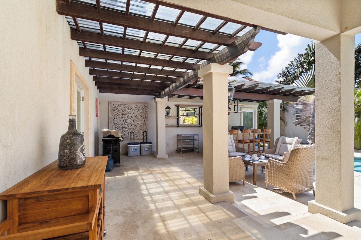 Un patio exterior con pérgola, mobiliario, piscina y zona de bar.