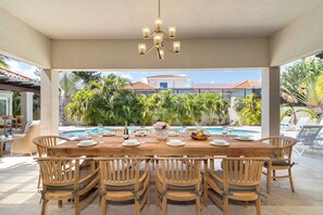 Zona d'esmorzar a l'aire lliure amb taula de fusta preparada per a sis persones, envoltada de cadires de vimetla, amb vista a una piscina i vegetació tropical.
