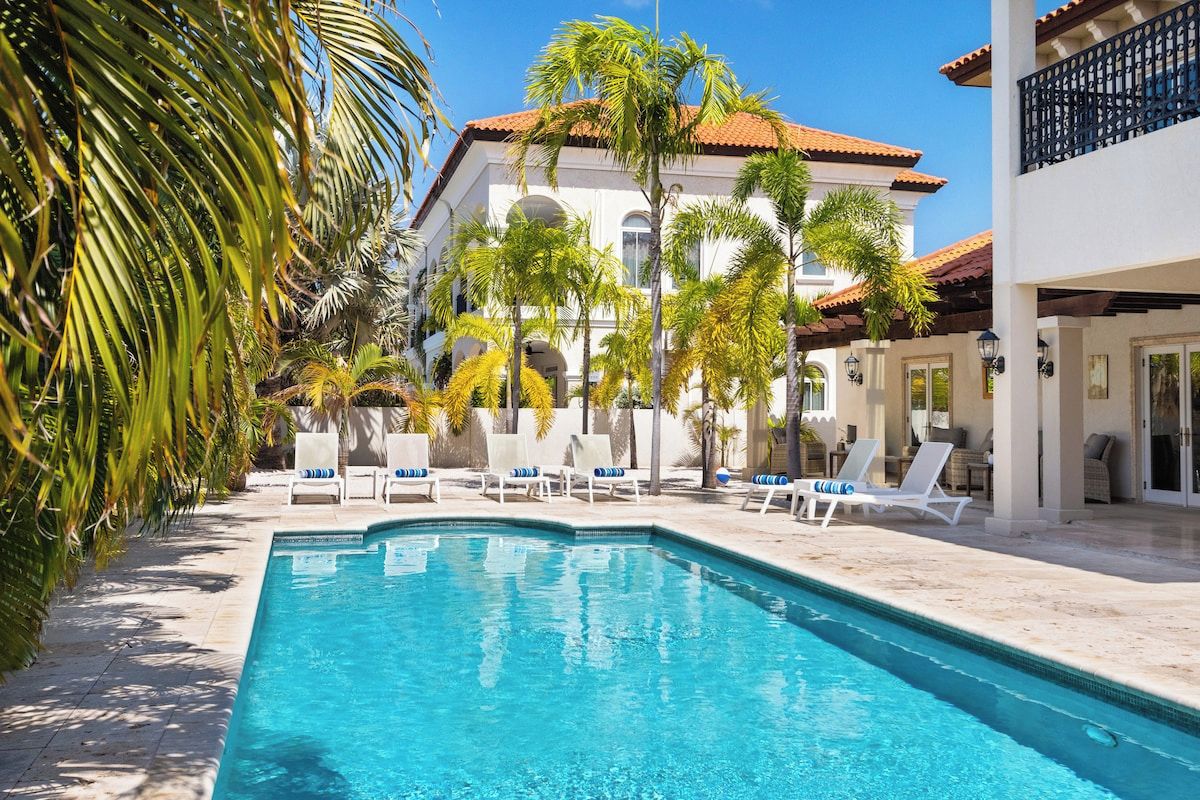 A swimming pool with lounge chairs, palm trees, and a building with a tiled roof.
