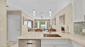A modern kitchen with a stainless steel dishwasher, white cabinets, a large island with a sink, pendant lights, and a view of the outdoors.