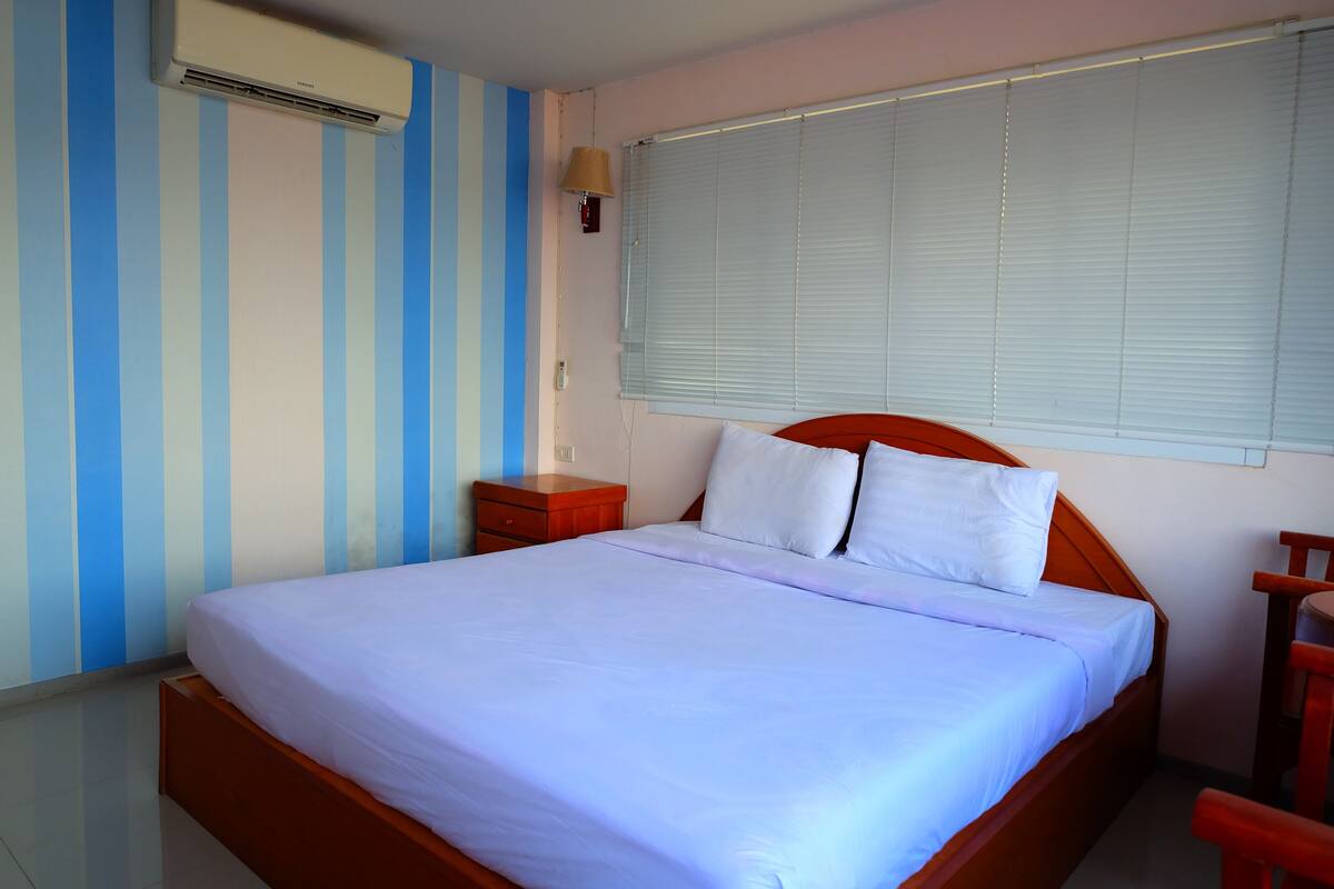 A neatly made bed with white bedding in a room with striped walls, a window with blinds, and a wooden nightstand.