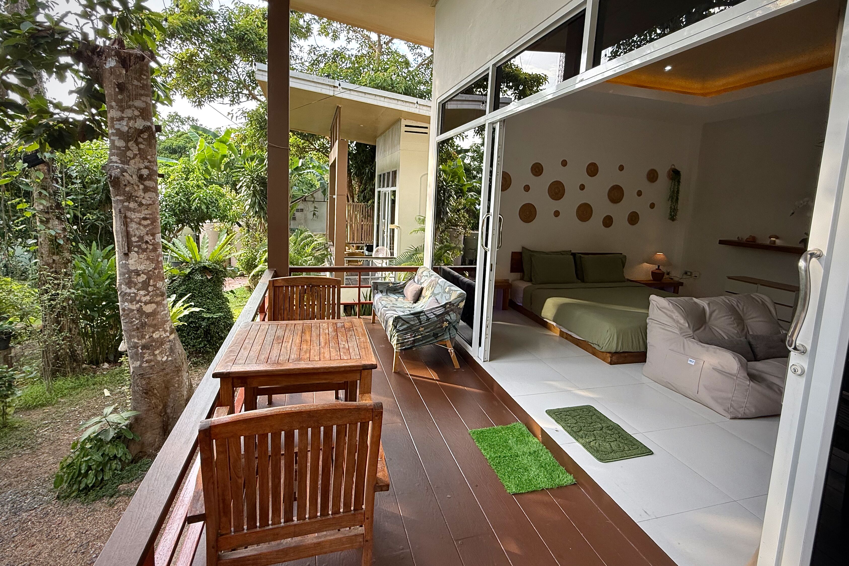 A modern outdoor patio with wooden furniture, a sofa, and a bed inside a room with a large glass door.