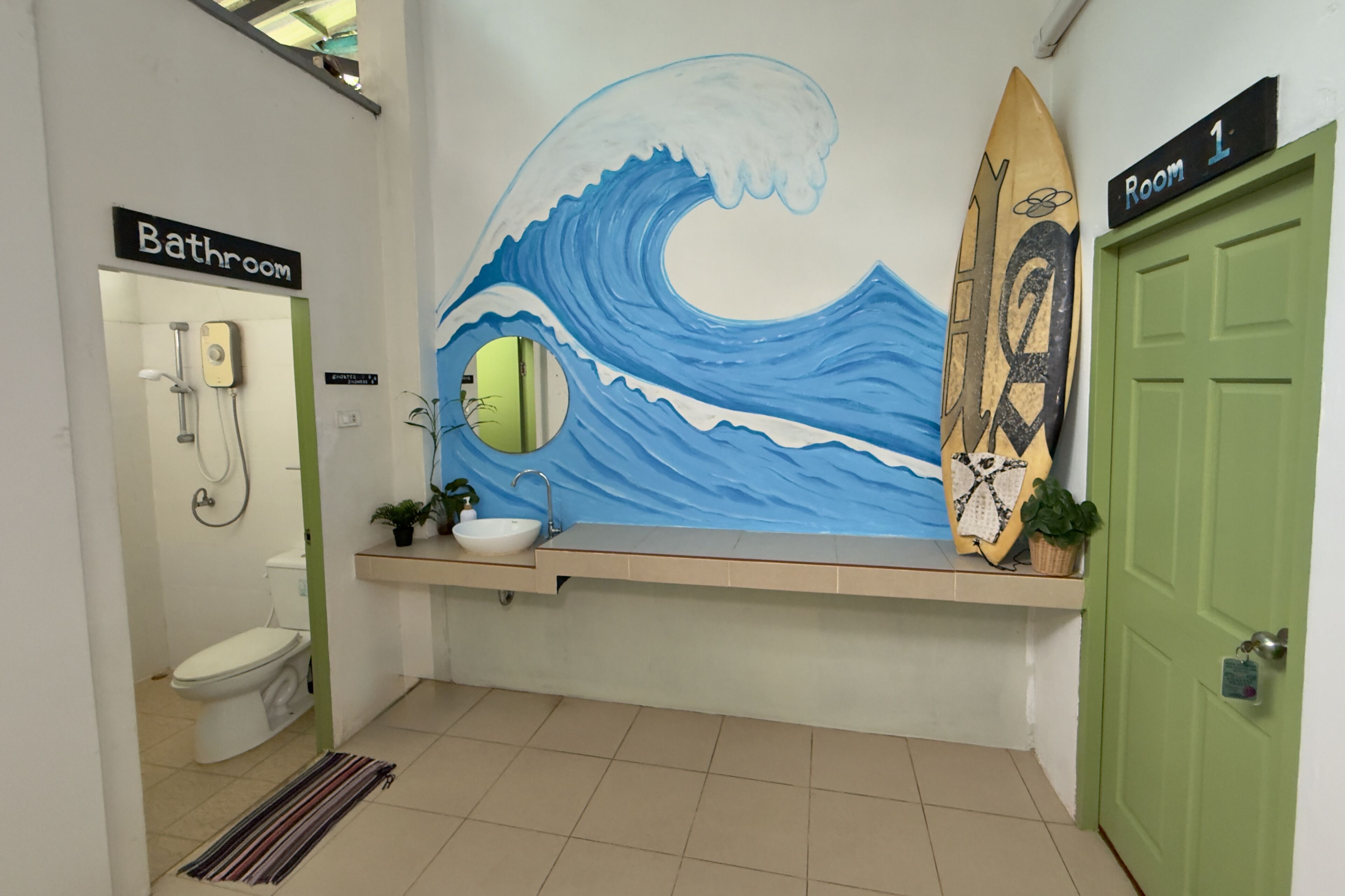 Baño con inodoro, lavabo y ducha. Se aprecia un mural con motivos de olas y una decoración con una tabla de surf.