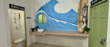 Baño con inodoro, lavabo y ducha. Se aprecia un mural con motivos de olas y una decoración con una tabla de surf.