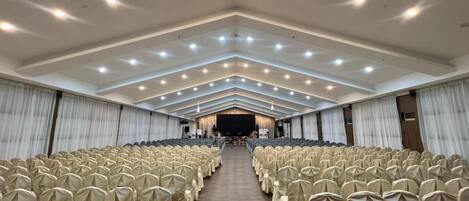 A large hall with rows of chairs covered in white cloth, a stage at the front, and a high ceiling with recessed lighting.