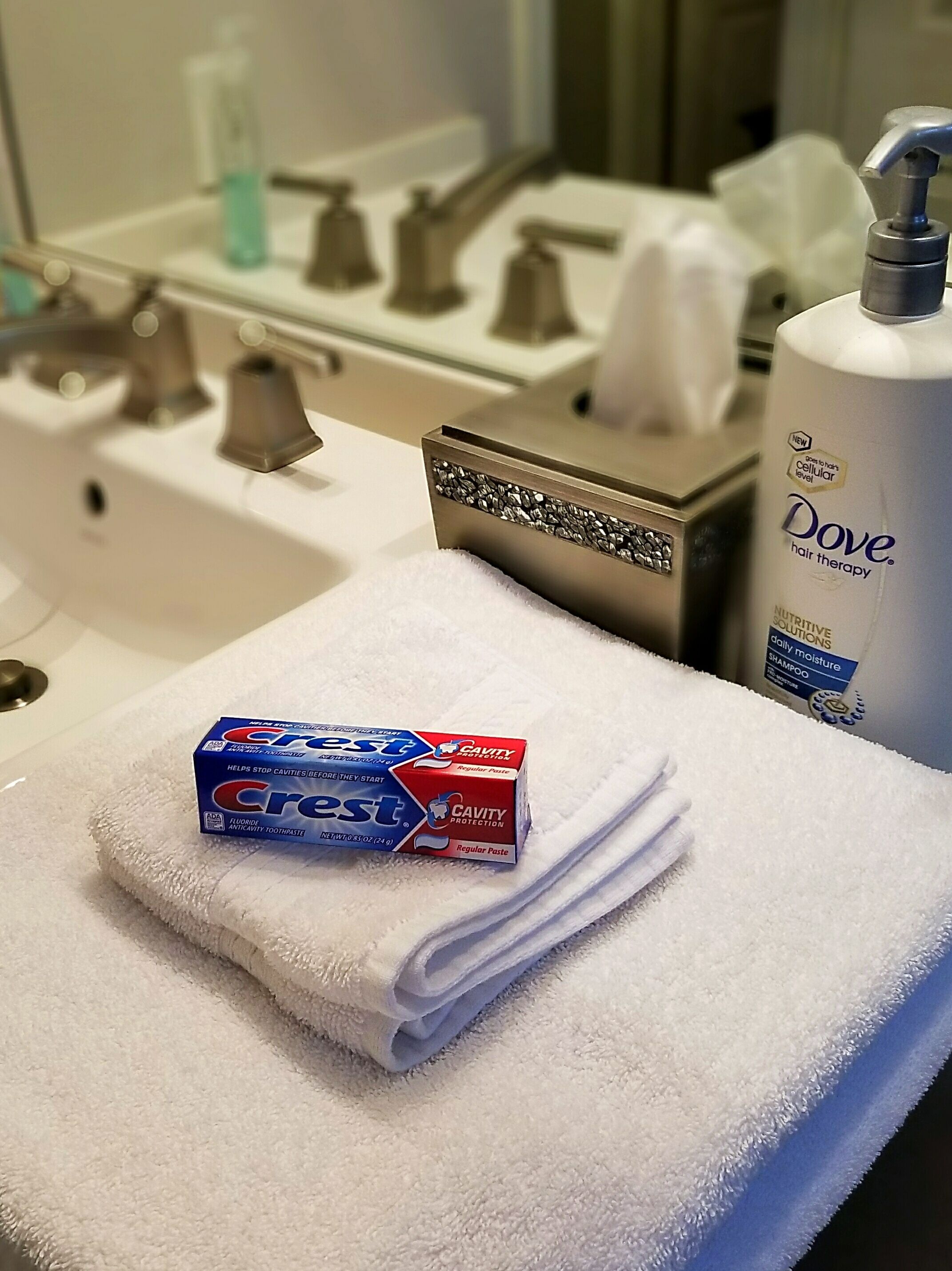 A bathroom with a white sink, a bottle of Dove hair therapy shampoo, and Crest toothpaste on a towel.